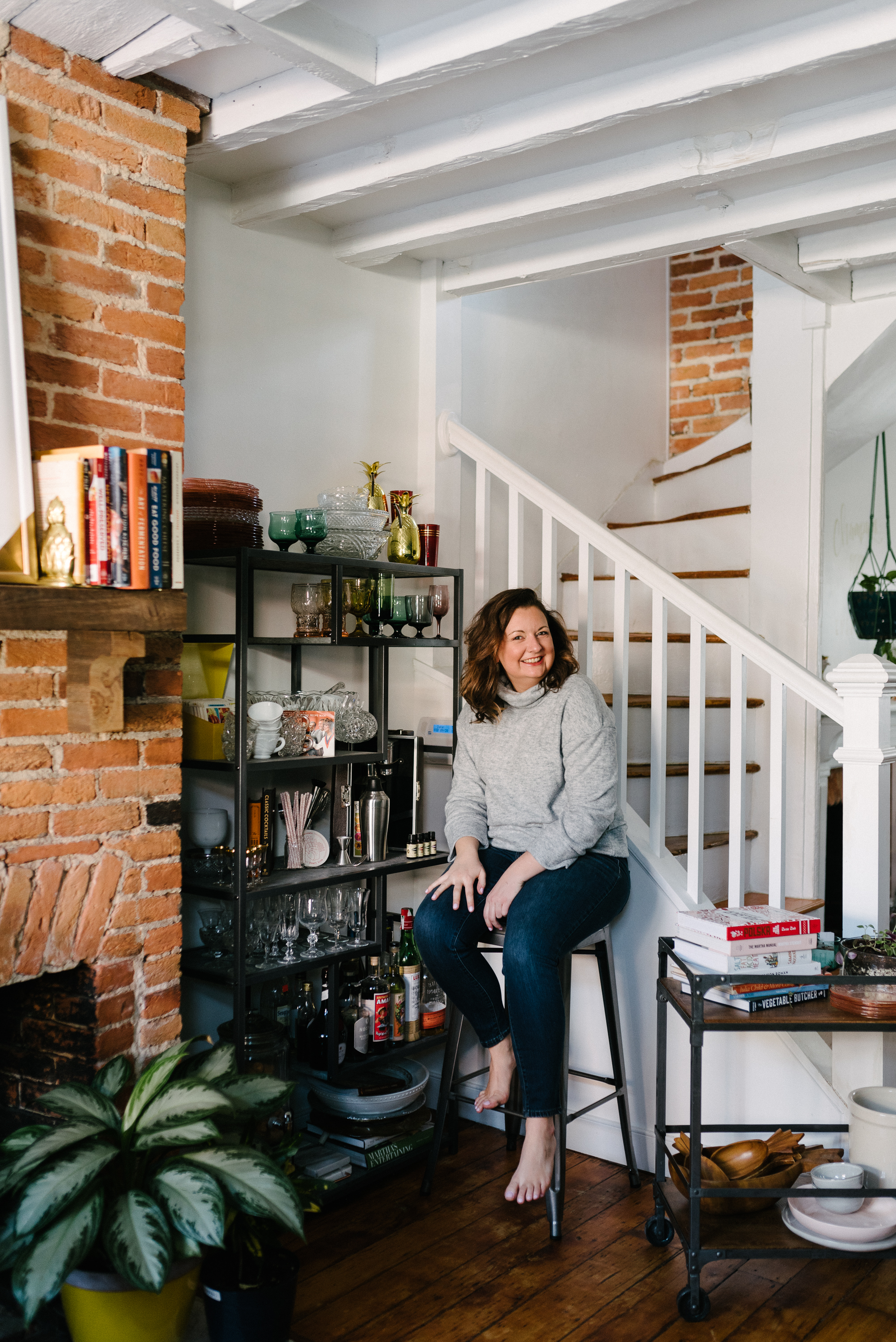 Kathleen sitting barefoot on a stool by a brick hearth, with a cart of cookbooks beside her