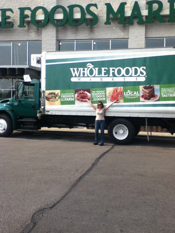 Kathleen standing with arms out in front of a Whole Foods Market refrigerated truck in a store parking lot