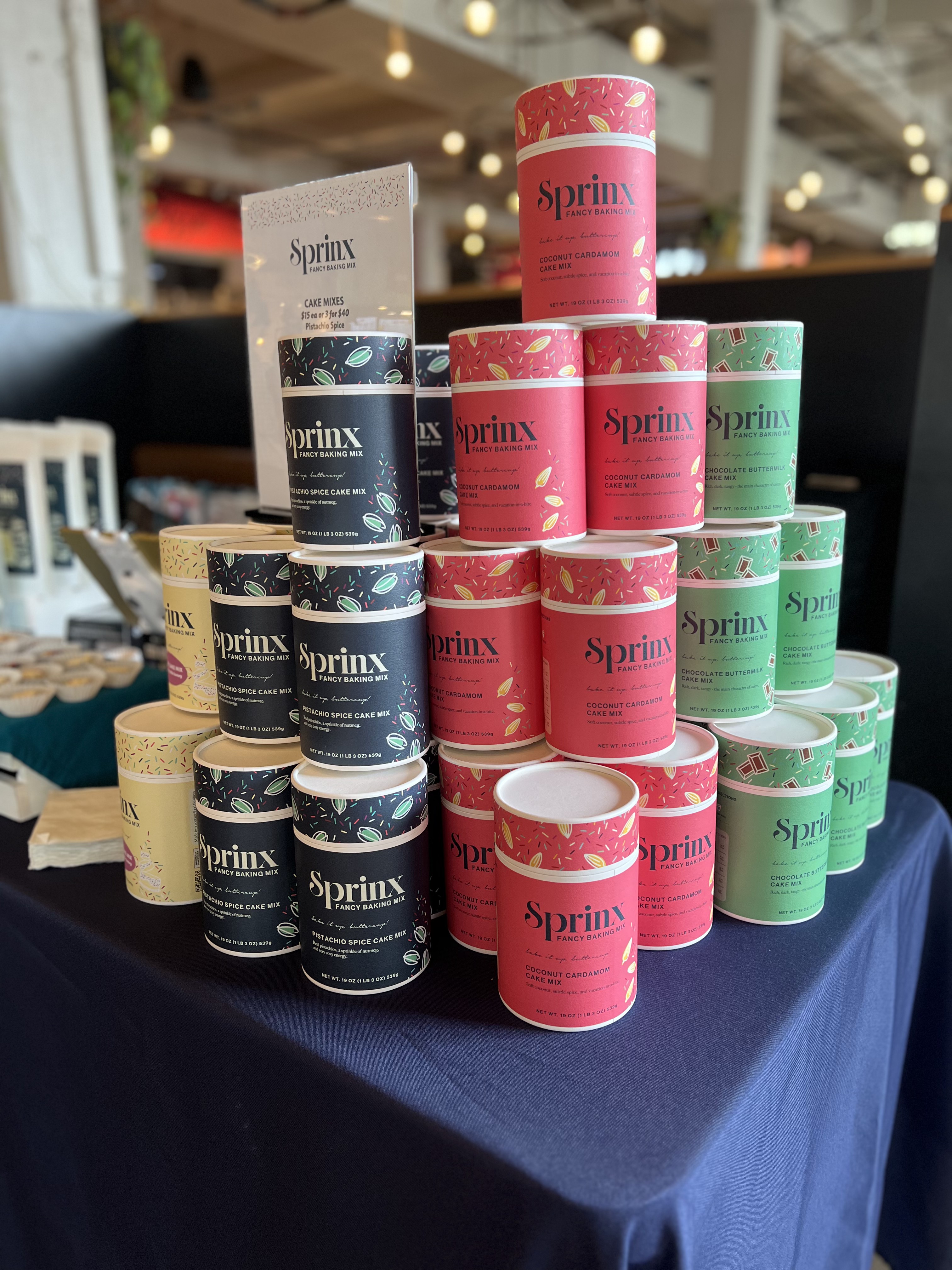 A pyramid of Sprinx Fancy Baking Mix canisters with custom sprinkle-patterned lids in pistachio spice, coconut cardamom and chocolate buttermilk