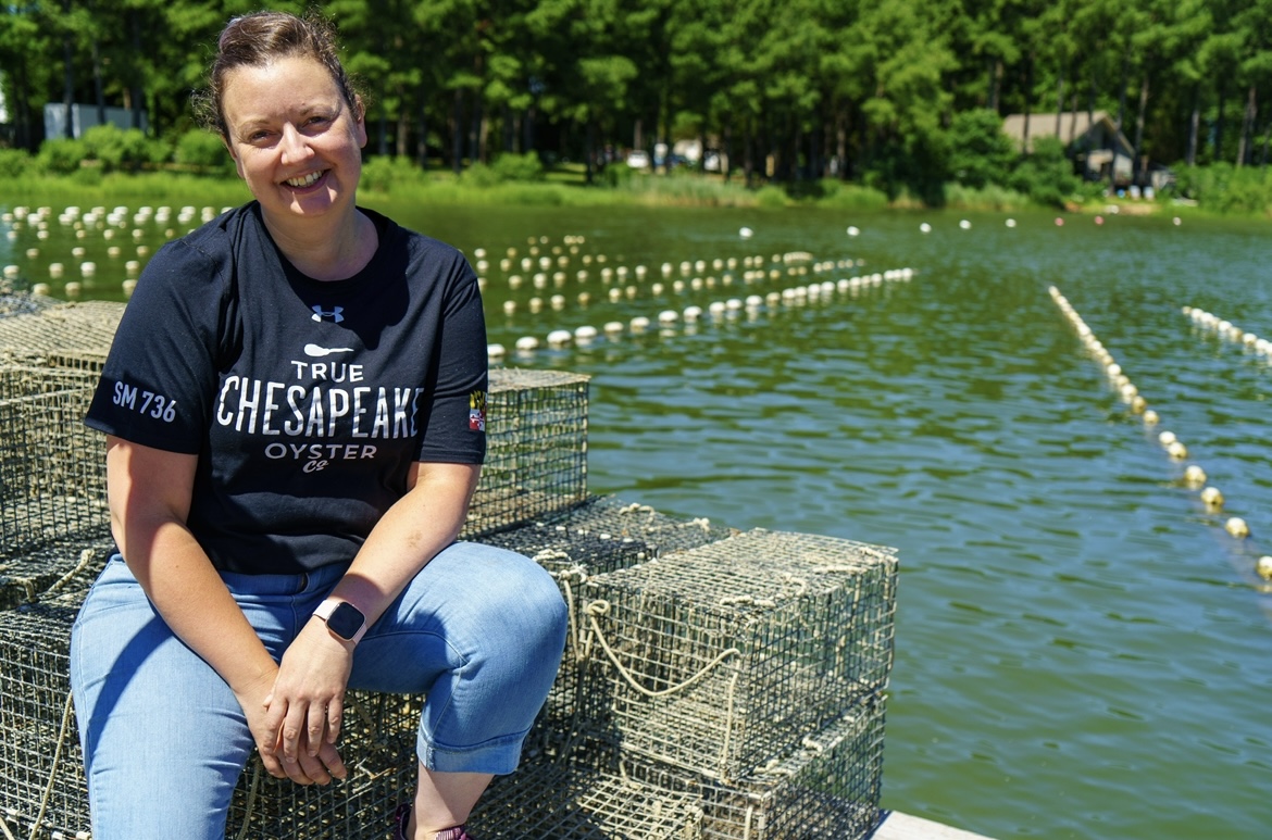 Kathleen sitting on stacked wire-mesh oyster cages with lines of buoys and a tree-lined shore behind her