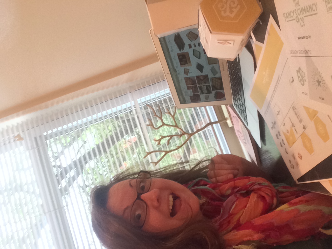 Kathleen laughing at a desk beside a laptop showing packaging artwork, with a hexagon box mockup