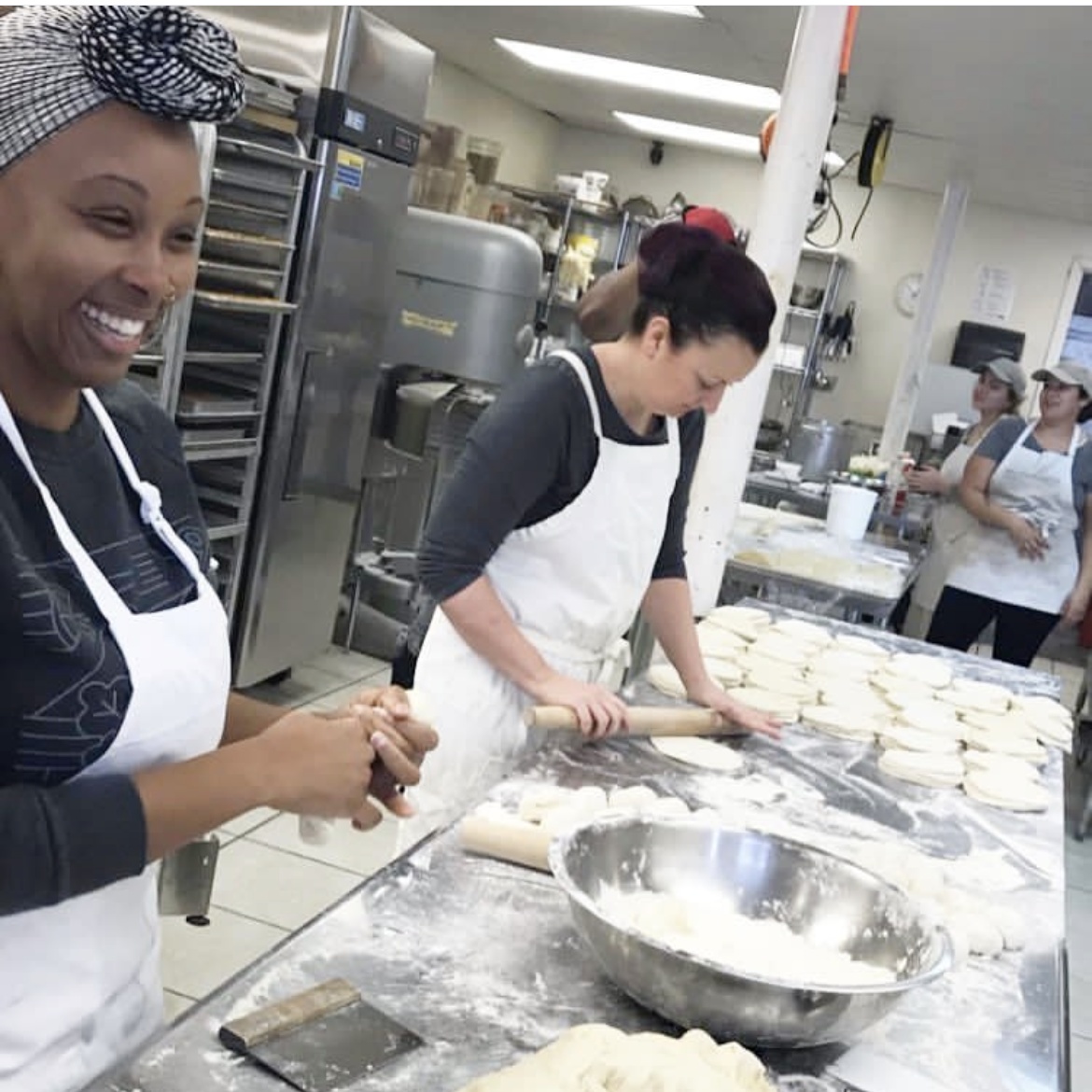 Kathleen laughing while shaping dough at a stainless-steel bakery table while a friend rolls out pita beside her