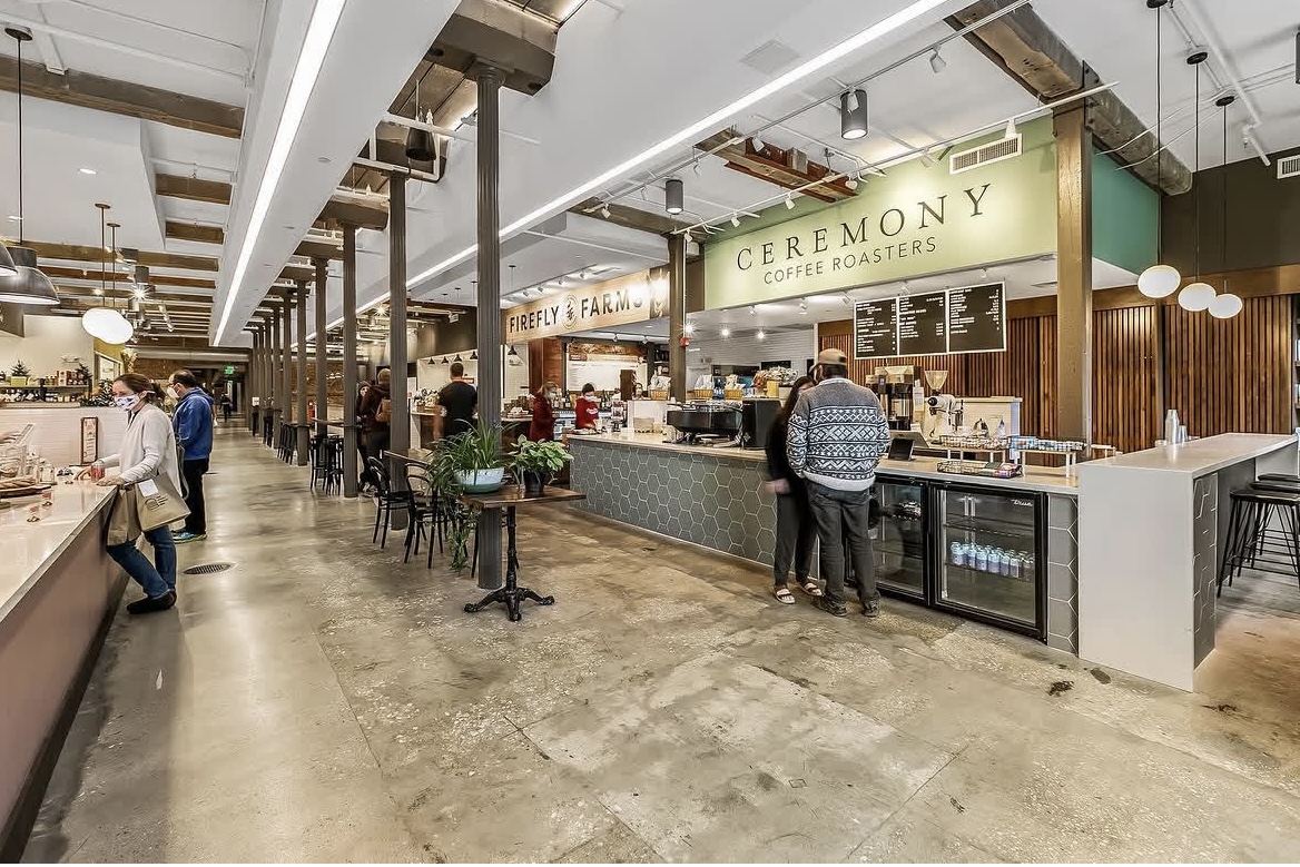 Inside the market: a long hall lined with vendor counters, including Ceremony Coffee Roasters and Firefly Farms, with structural columns, pendant lights, and shoppers walking through