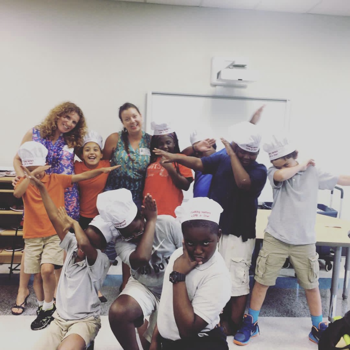 Kathleen standing with a fellow instructor beside a classroom of children wearing paper Cooking Matters chef hats, several striking playful poses