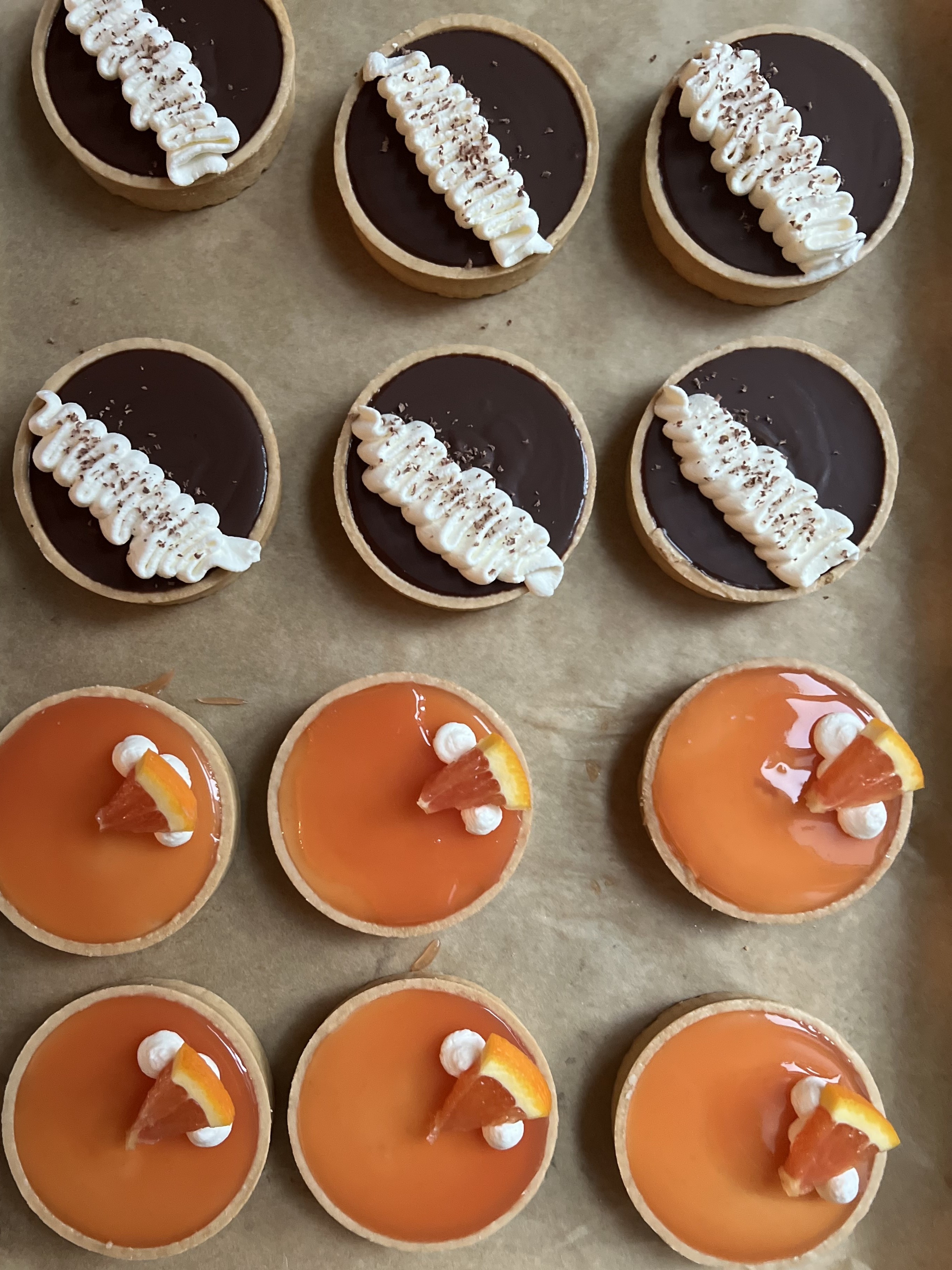 A sheet pan lined with parchment holding twelve tarts: six dark chocolate ganache tarts topped with ruffled piped cream and chocolate specks, and six glossy blood-orange curd tarts topped with cream dollops and citrus wedges
