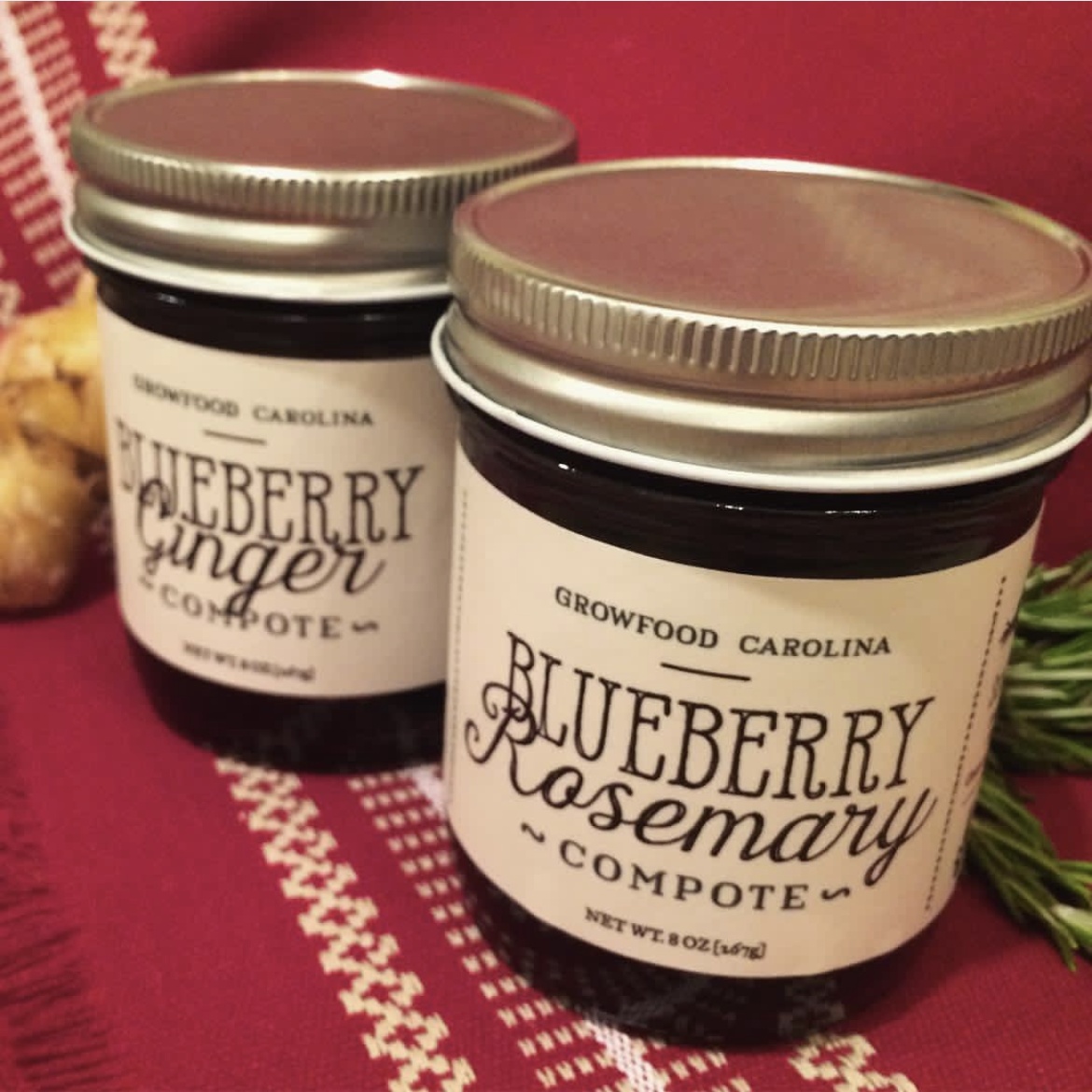 Two glass jars of GrowFood Carolina compote, Blueberry Rosemary and Blueberry Ginger, on a burgundy woven cloth with rosemary sprigs and a piece of ginger root